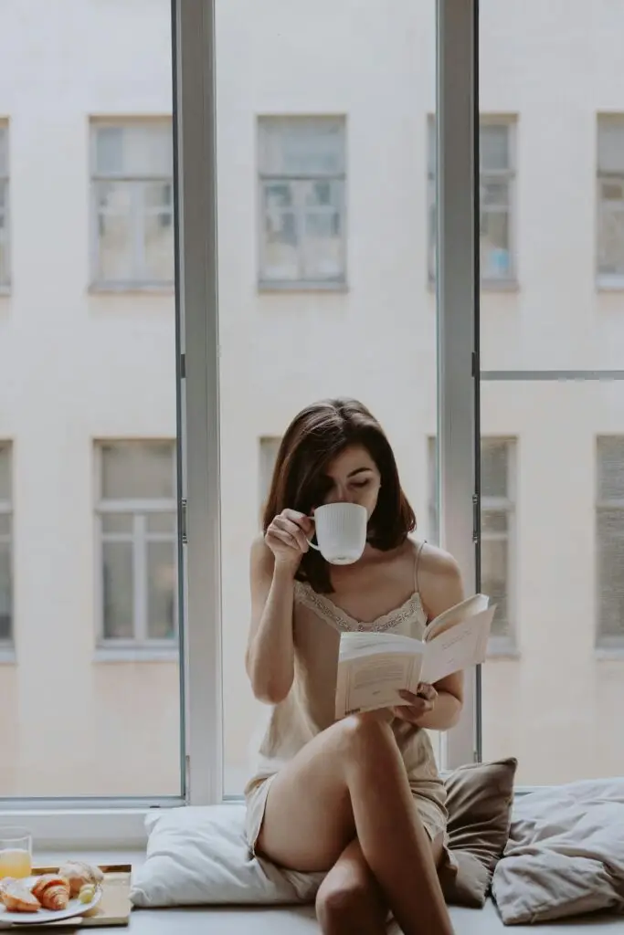 A woman relaxes with a book and coffee by a window in an urban setting, exuding a peaceful morning vibe.